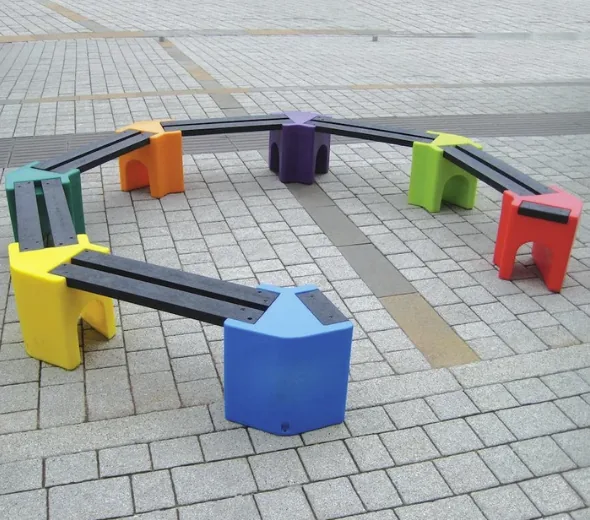 A multi-coloured plastic and recycled-board bench curved into a horseshoe shape on a paved surface