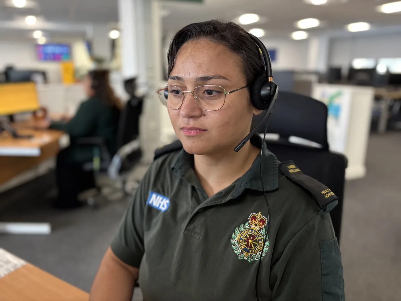 A London Ambulance Service 111 health adviser in uniform, glasses and a headset at her desk in a control room