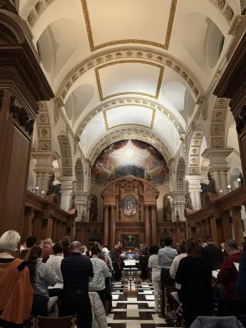 A congregation standing in the nave of St Bride's Church, with an arched white and gold ceiling and a painted screen behind the altar