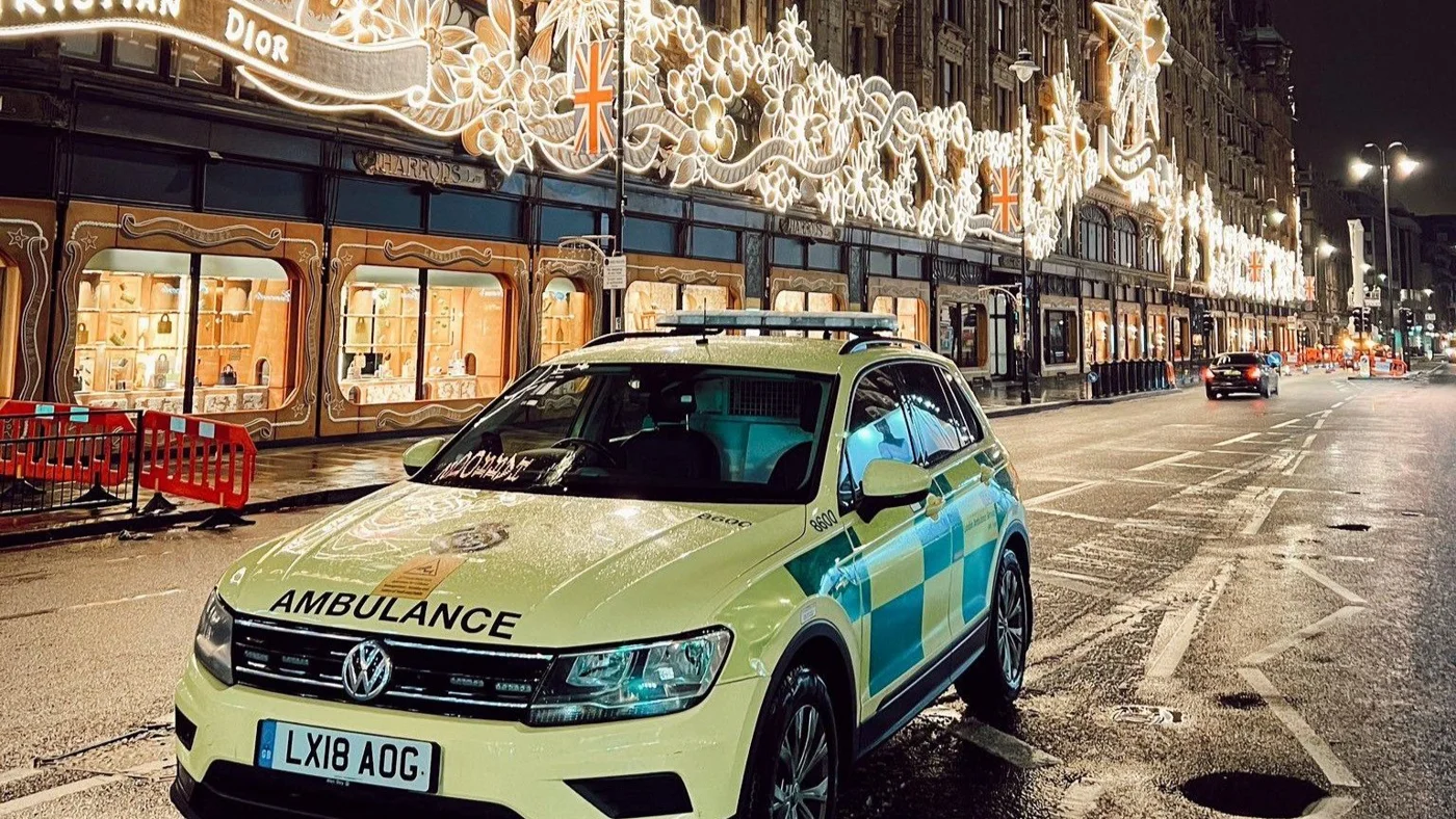 A London Ambulance Service response car parked at night on a wet street below shop fronts hung with Christmas lights