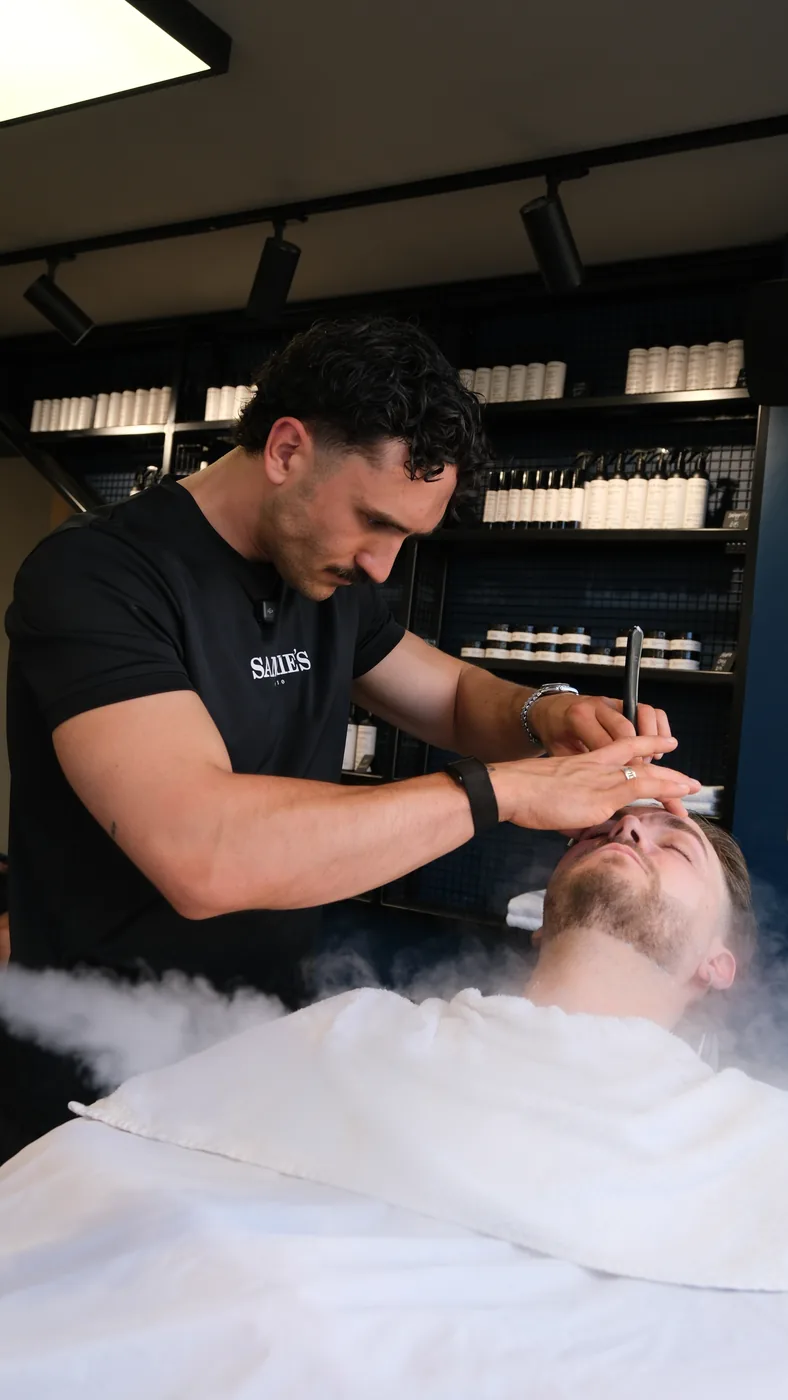 A barber gives a customer a straight-razor shave beside a shelf of grooming products