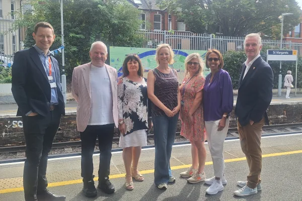 Seven people standing together on the platform at St Margarets station, in front of a painted mural on the far side of the tracks.