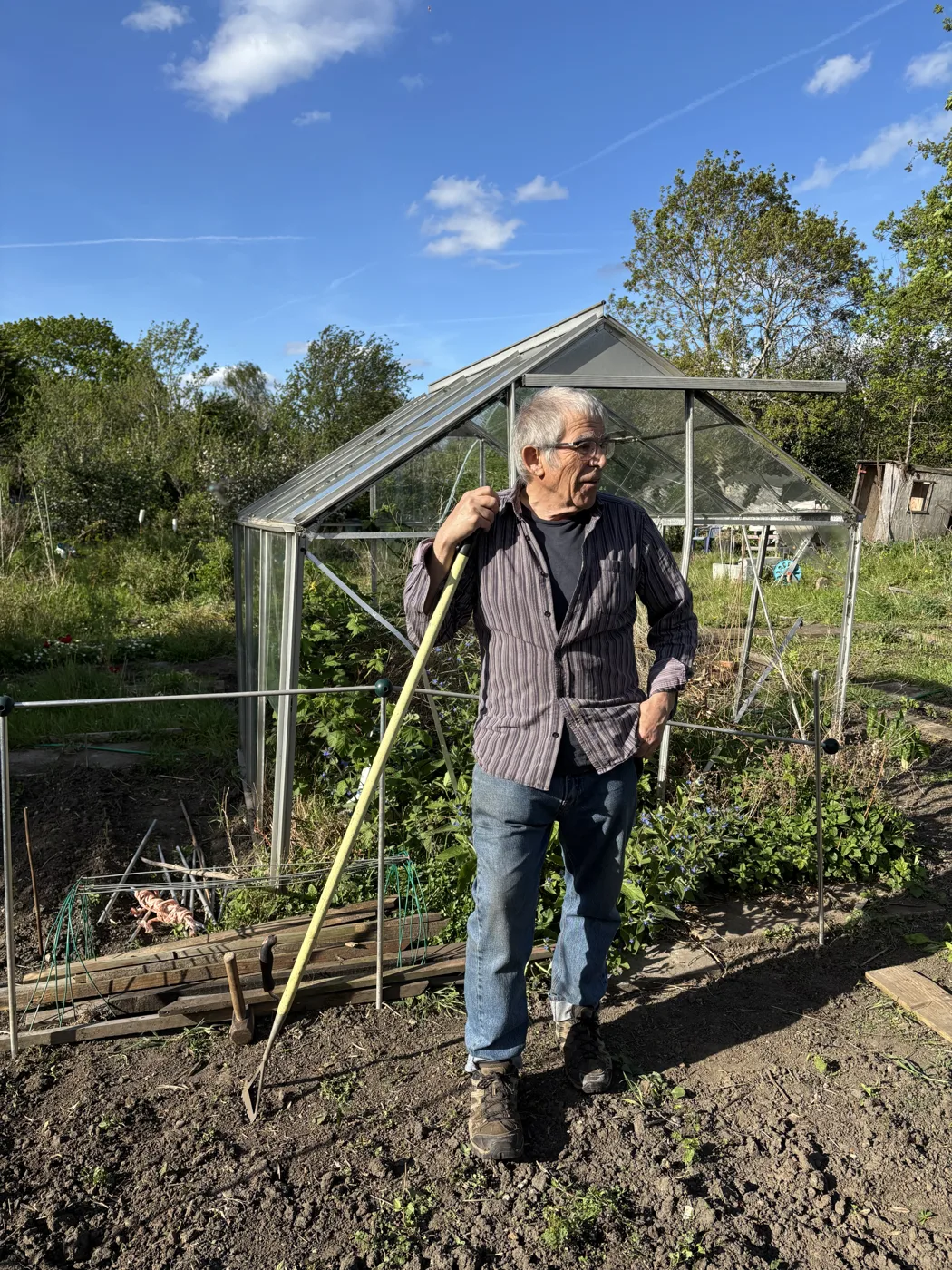 A plot holder with a hoe standing in front of a greenhouse on an allotment plot